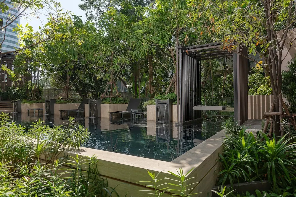 A pool surrounded by trees and plants, with dark sun loungers and a metal pergola beside the water.
