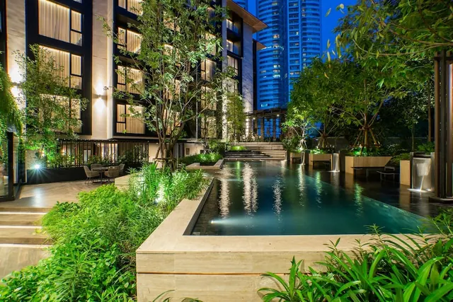 A long outdoor pool at night is lined with tropical plants and trees, with a hotel building on the left and an illuminated high-rise tower behind.