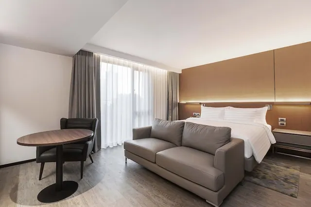 A hotel bedroom with a king bed, a grey sofa, a round wooden table with a dark chair, and sheer curtains over a large window.