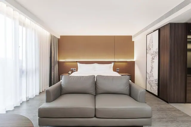 A bedroom with a made double bed behind a grey leather sofa, sheer curtains on the left and a wood-panelled wardrobe on the right.