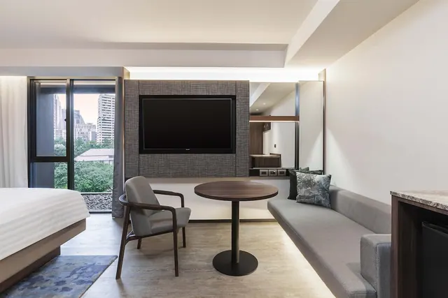 A hotel room with a bed at left, a wall-mounted television, a grey sofa, a round wooden table with a chair, and a window overlooking high-rise buildings and trees.