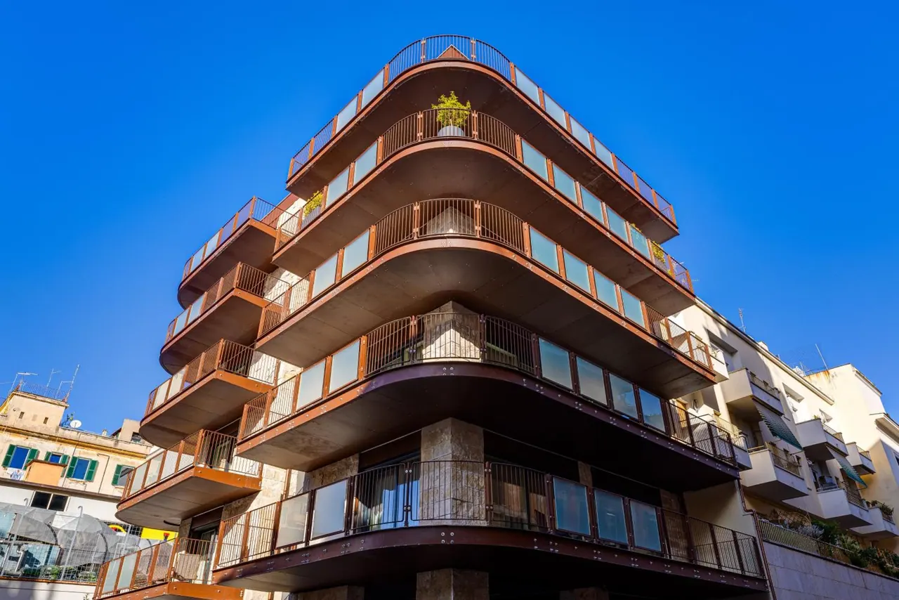 A curved multi-storey building with rust-coloured balconies and glass balustrades rises against a clear blue sky.