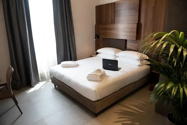 A made double bed with white linen, folded towels and a laptop on it, beside a wood panelled headboard and a window with grey curtains.
