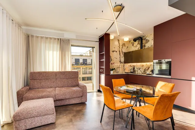 A living area with a sofa, a dining table with orange chairs, a built-in kitchen, and a glass door open to a balcony.