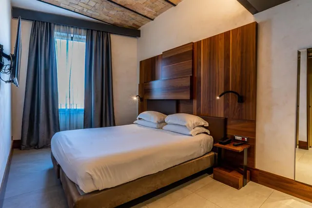A double bed with white linen and a wooden headboard panel stands in a bedroom with a brick vaulted ceiling, grey curtains and a wall-mounted television.
