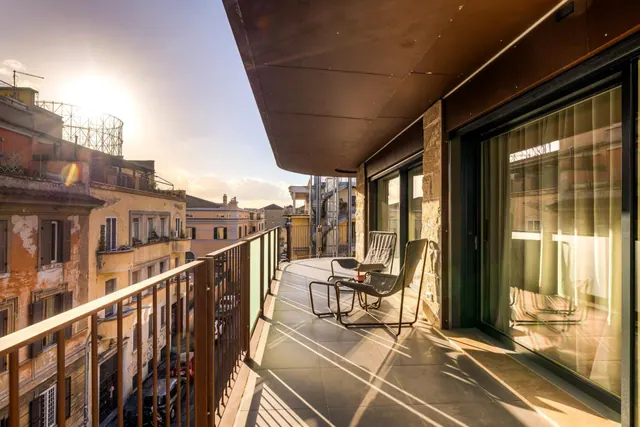 A balcony with two metal chairs and a small table overlooks sunlit Roman buildings at sunset, with glass sliding doors leading inside.
