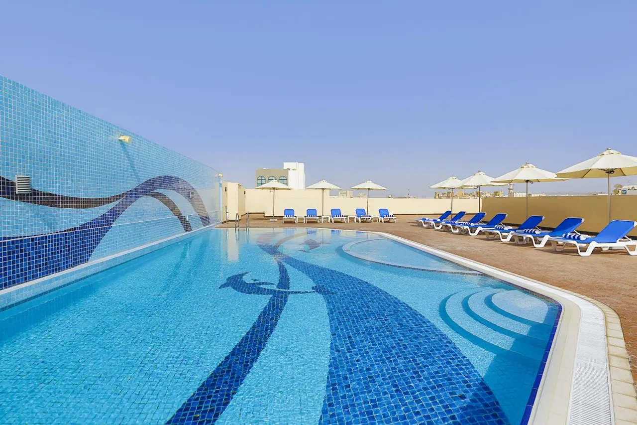 A rooftop swimming pool with blue mosaic tile designs, surrounded by blue sun loungers and beige umbrellas under a clear sky.