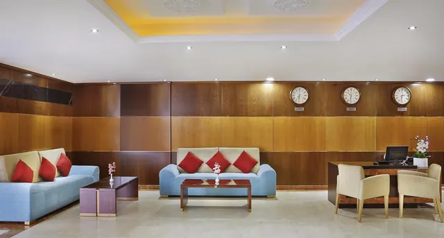 A hotel lobby with wood-panelled walls, light blue sofas with red cushions, a coffee table, a reception desk with a computer monitor, and three wall clocks.