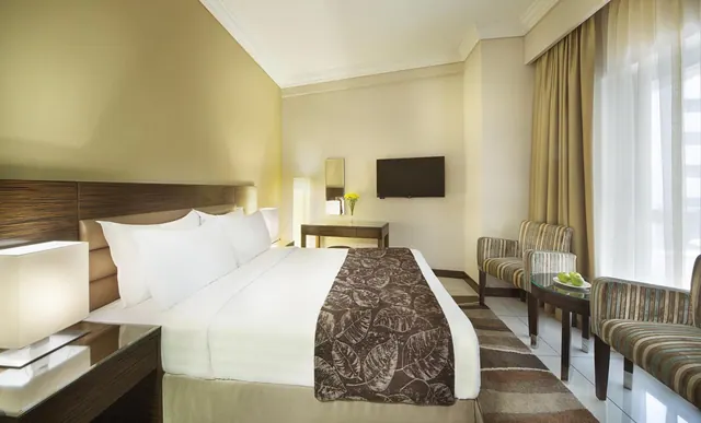 A bedroom with a made double bed, a wall-mounted television, a small desk, and striped armchairs beside a window with sheer and patterned curtains.