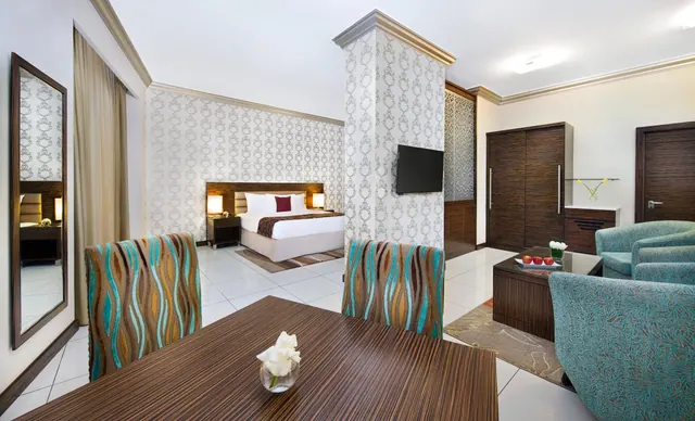 A hotel suite with a wooden dining table in the foreground, teal armchairs and a coffee table on the right, and a double bed with a wall-mounted television visible in the background.