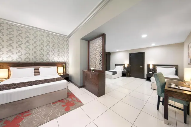 A hotel bedroom with a double bed on the left, two single beds in the background, a decorative partition screen and a wooden desk with a chair on the right.