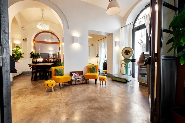 A lobby lounge with arched ceilings, yellow armchairs, small yellow stools, a vintage scale and a dining table in the background.