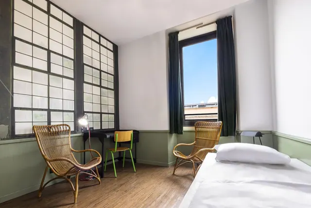 A bedroom with a white-made bed, two rattan armchairs, a black desk with a green chair, and tall grid-paned windows, with a further window showing blue sky.