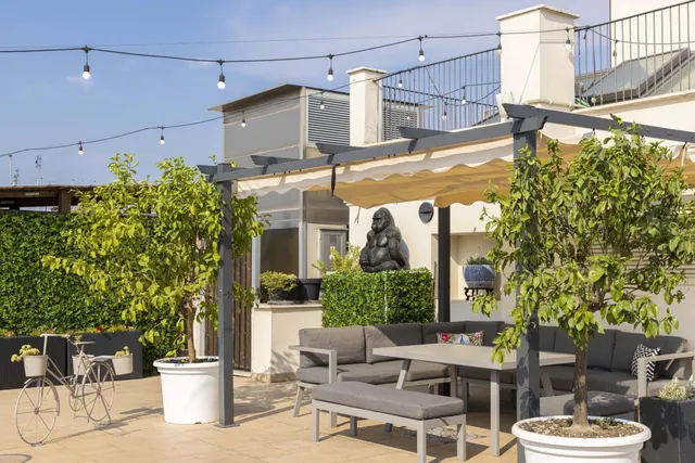 A rooftop terrace with a pergola, string lights, grey sofas, a dining table and potted lemon trees, with a gorilla sculpture and a hedge behind.