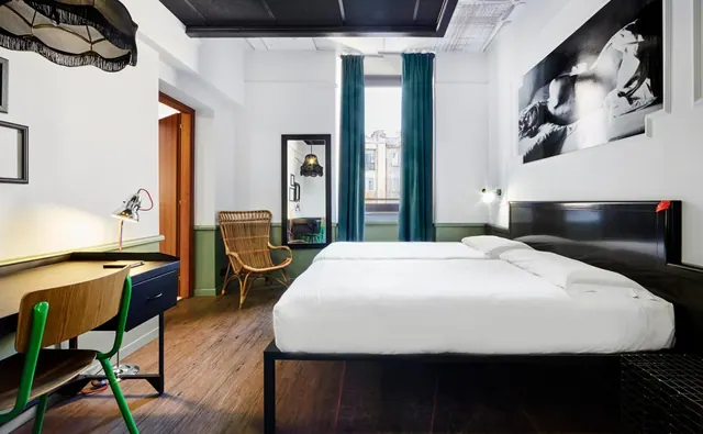 A bedroom with a made double bed under a large black and white photograph, a wooden desk with a green chair, and a wicker armchair beside a window with teal curtains.