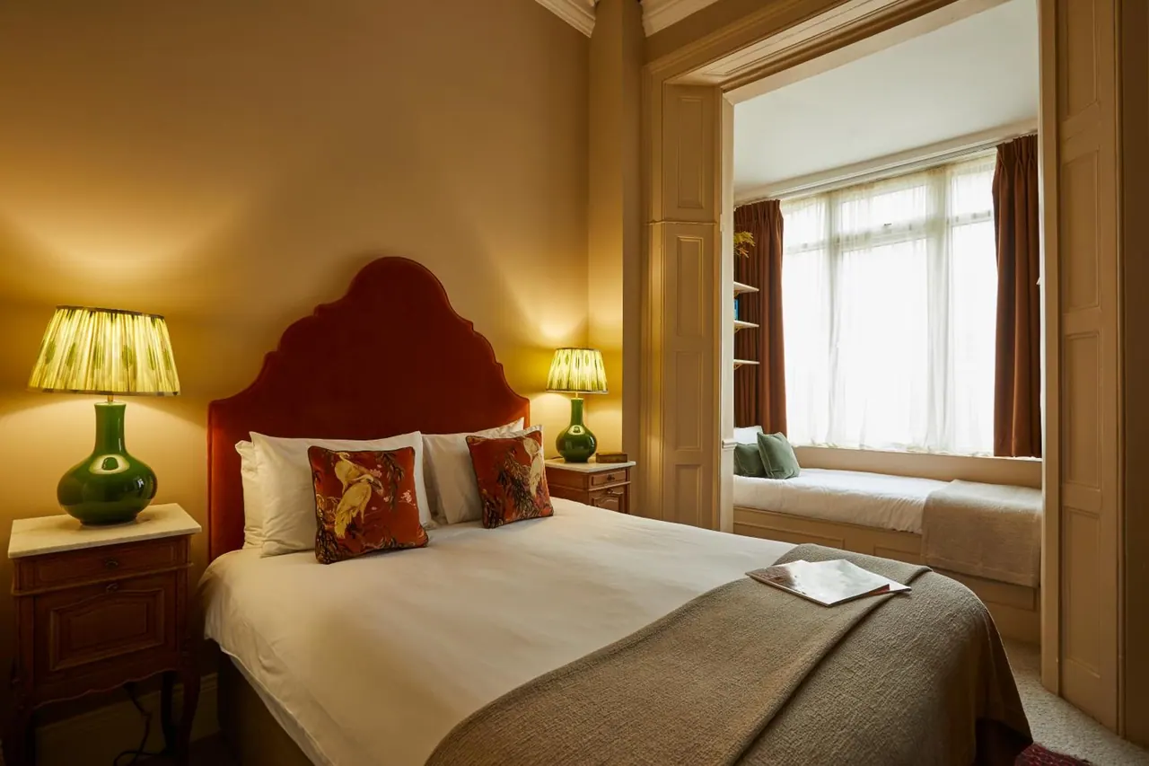 A double bed with a red velvet headboard and green table lamps on each side, with a bay window and window seat visible in the adjoining alcove.