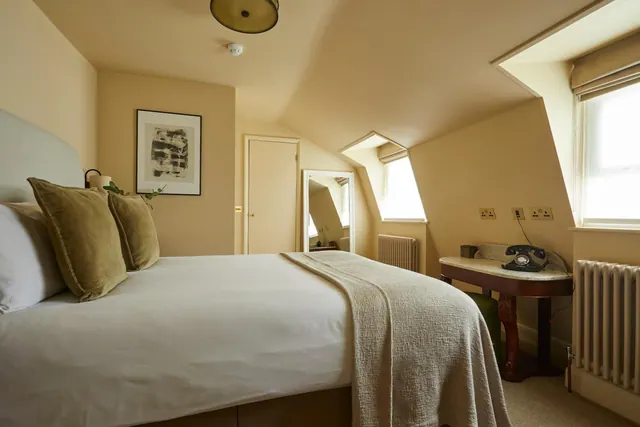 A bedroom with a double bed, olive cushions, a marble-topped console table with a black telephone, and sloped ceilings with dormer windows.