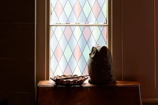 A carved wooden owl figurine and a carved wooden dish sit on a wooden chest in front of a window with pastel diamond-patterned leaded glass.