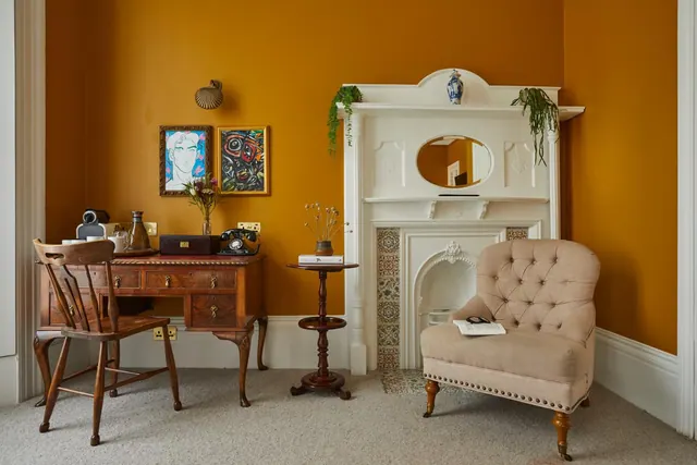 A room with mustard walls holds a wooden writing desk and chair, a white fireplace with a mirror, and a beige tufted armchair.