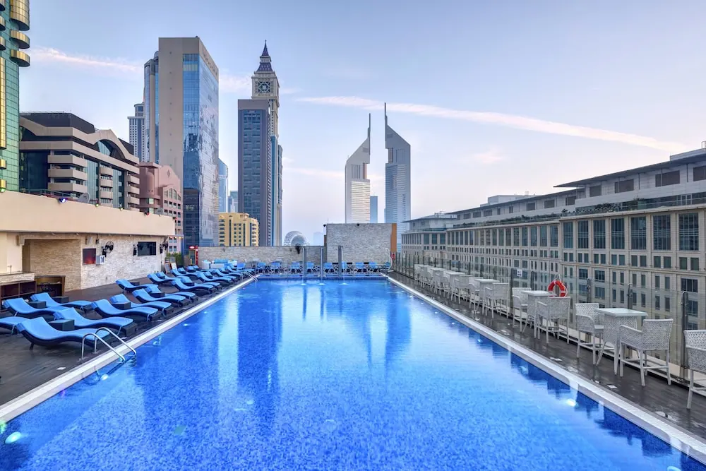 A rooftop swimming pool with blue loungers and white outdoor chairs, with city towers and the Emirates Towers in the background.