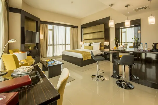 A staged hotel room with a double bed, yellow armchairs, a glass-topped desk, a bar counter with stools and a wall-mounted television.