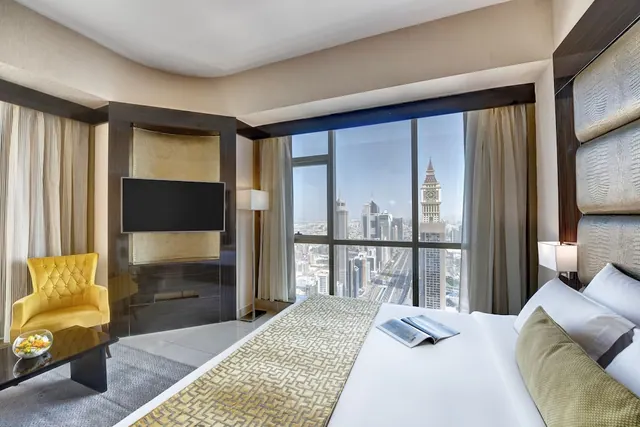 A bedroom with a made bed, a yellow armchair, a wall-mounted television and large windows overlooking a city skyline with a clock tower.