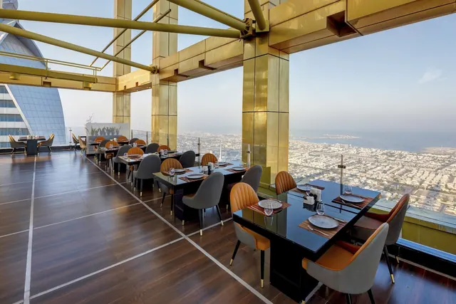A restaurant dining room with set tables and gold-framed columns, with floor-to-ceiling windows overlooking a city and the sea.