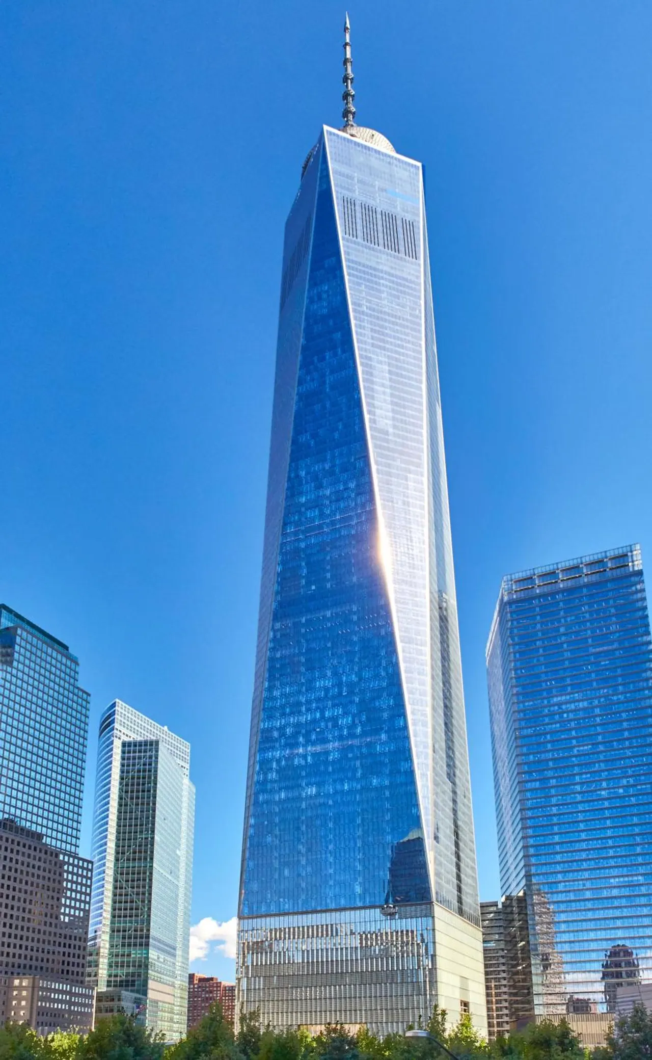 A tall glass skyscraper, One World Trade Center, rises against a clear blue sky with two other glass towers on either side and trees at its base.