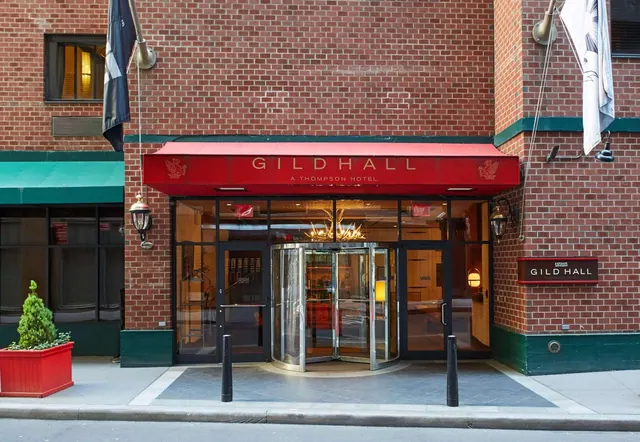The street-level entrance of Gild Hall, a Thompson Hotel, with a red awning over revolving doors and a brick facade.