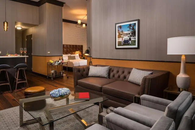 A hotel suite living area with a tufted brown leather sofa, grey armchairs, a glass coffee table, and a bed visible in the adjoining room.
