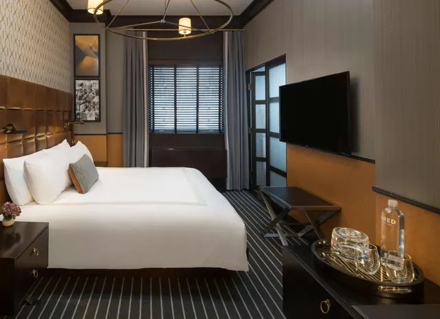 A bedroom with a made king bed, a wall-mounted television, and a tray of glasses and a water bottle on a dark dresser.