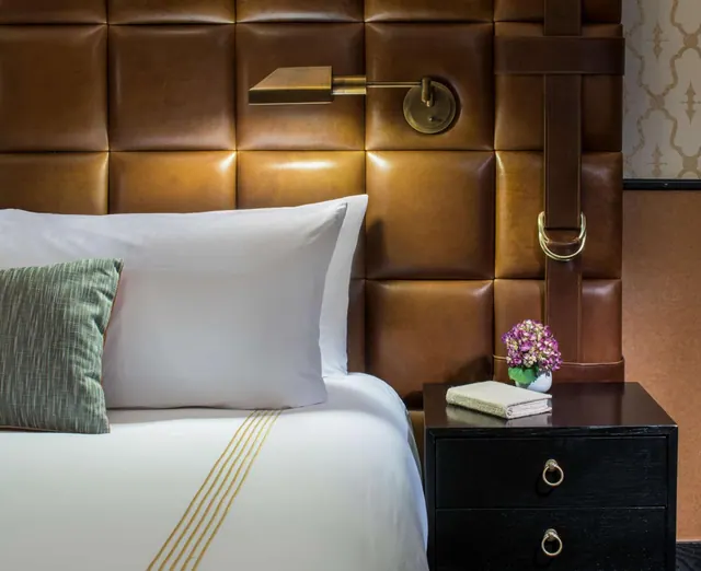 A made bed with a tufted leather headboard, a brass wall reading lamp, and a black nightstand holding a small flower vase and folded towel.