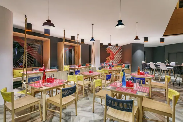 A modern restaurant dining room is arranged with wooden tables, colorful chairs, pendant lights and wall seating.