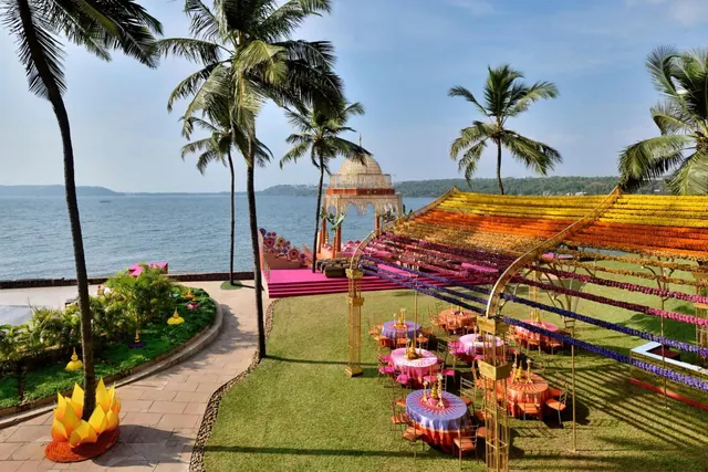 A decorated seaside garden is arranged with colorful event canopies, round dining tables, palm trees and a domed ceremony structure.