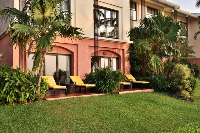 A tropical hotel building is shown beside a landscaped lawn with palm plants and outdoor loungers.