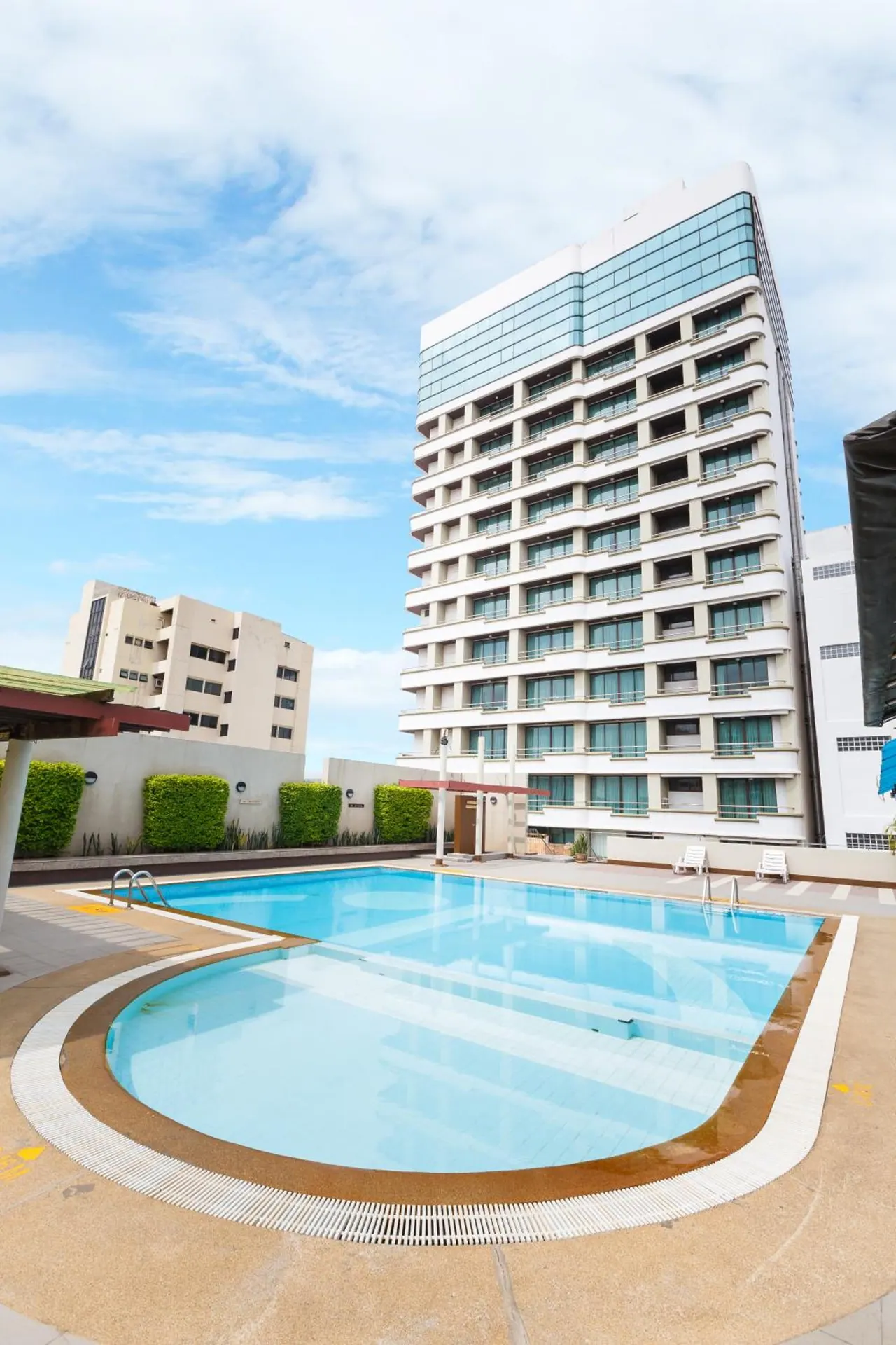 An outdoor swimming pool with a curved edge and two white sun loungers sits in front of a tall white hotel tower under a partly cloudy sky.