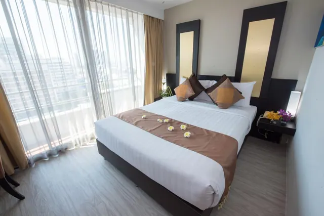 A made double bed with a brown runner and frangipani flowers faces floor-to-ceiling windows with sheer curtains and a view of city buildings.