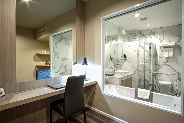 A wooden wall-mounted desk with a dark upholstered chair and black table lamp sits beside a glass-walled bathroom with marble tiles, a bathtub and a walk-in glass shower.