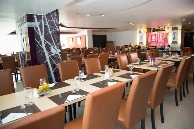 A dining room with rows of set tables and brown leather chairs, a dark pillar with a white branch pattern on the left, and a pink Japanese-style food counter at the back.
