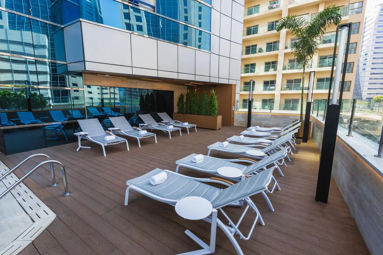 Rows of white sun loungers with folded white towels and round side tables line a wooden pool deck beside a glass-fronted building.