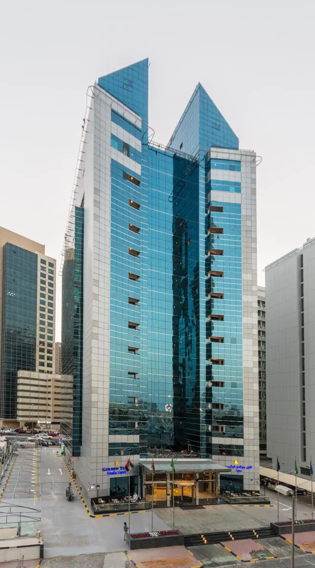 The photo shows the tall glass-fronted Golden Tulip tower with two pointed tops, with its entrance canopy and signage at street level, a parking area and neighbouring buildings beside it.