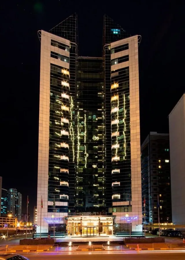 Night exterior of a tall glass and stone hotel tower with the Golden Tulip Media Hotel sign above its lit entrance canopy, with cars and flags visible at street level.