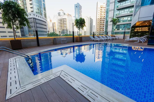 An outdoor pool with blue tiles, white sun loungers on the deck, and high-rise towers visible beyond the wall.