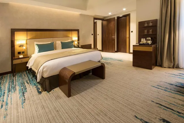 A hotel bedroom with a made king bed, a padded bench at the foot of the bed, bedside lamps, and a wooden console holding a kettle and glasses.