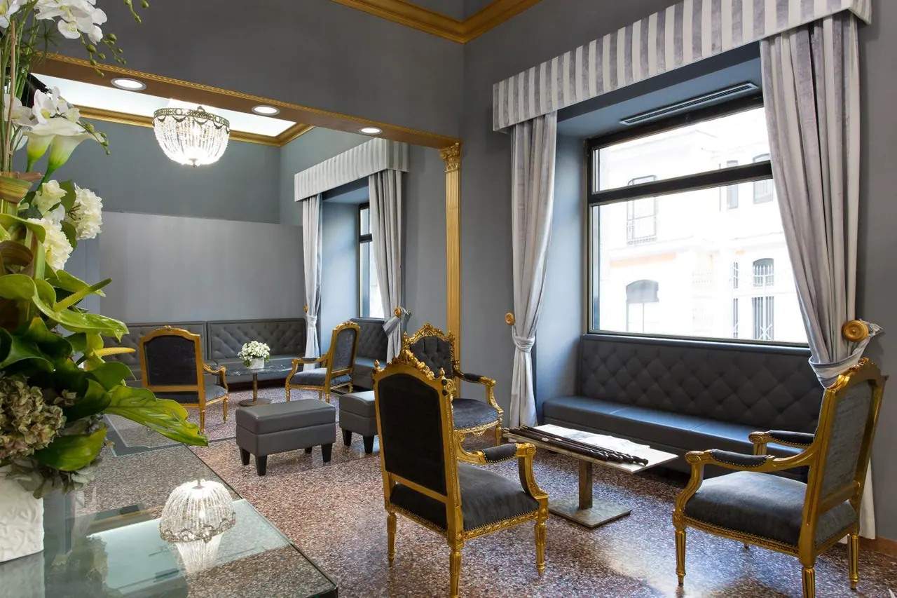A lounge seating area with gilded armchairs, grey upholstered sofas, a glass coffee table and a crystal chandelier, with a window onto a neighbouring building facade.