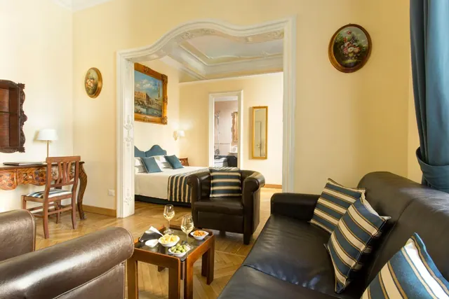 A hotel suite living room with a leather sofa and armchair, a coffee table with wine glasses, and an arched doorway leading to a bedroom with a double bed.