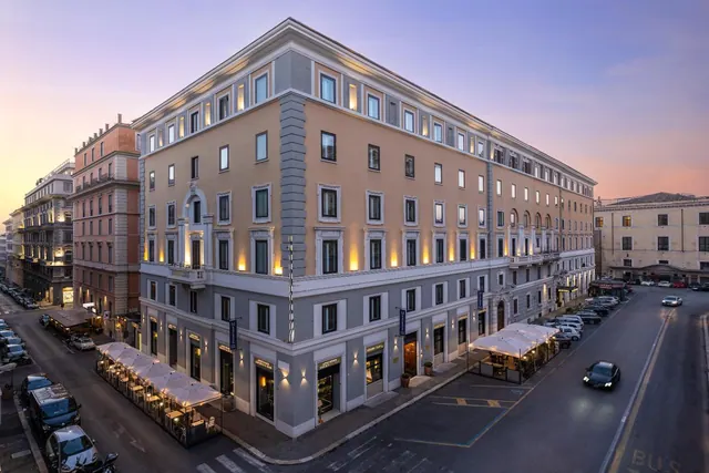 Exterior of a cream-coloured multi-storey hotel building at dusk, with lit windows, outdoor café tables under white umbrellas, and cars on the street.