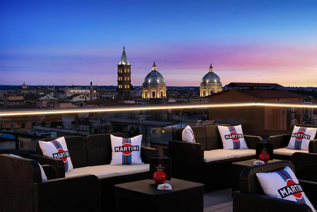 A rooftop terrace with wicker sofas and Martini-branded cushions overlooks city rooftops, church domes and a bell tower at dusk.
