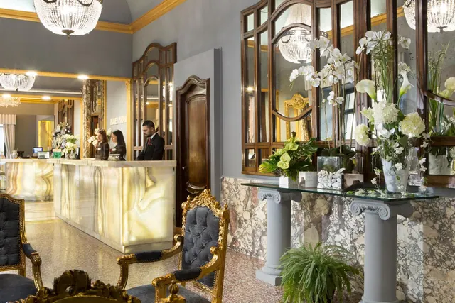 A hotel lobby with an illuminated onyx reception desk, gold-framed chairs, mirrored wall panels, a marble console table with white flowers, and two staff members at the desk.