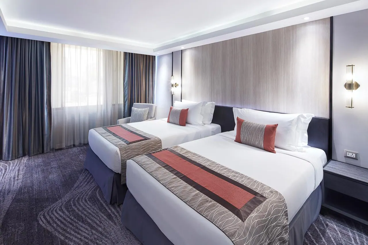 A bedroom with two beds with red and grey runners, an armchair by a window, and blue curtains.